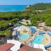 Complesso acquatico con piscine, ombrelloni blu e vista sul mare e colline verdi.