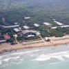 Spiaggia con bagnanti e ombrelloni, circondata da vegetazione fitta e alcuni edifici.